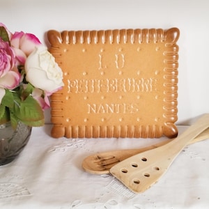 Peut inclure: Une plaque décorative en forme de biscuit avec les mots "LE PETIT BEURRE NANTES" en relief sur sa surface. La plaque est marron clair avec un bord strié. Des ustensiles de cuisine en bois et un vase de fleurs roses et blanches sont également présents.