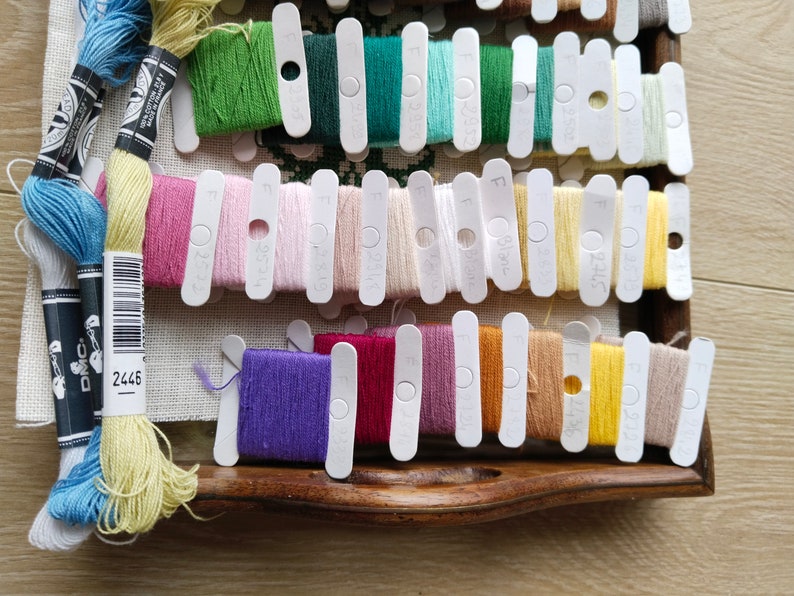 May include: A close-up of a wooden box filled with colorful embroidery floss spools. The spools are organized by color and labeled with DMC numbers. The box is open and the spools are visible. The DMC number 2445 is visible on the box.