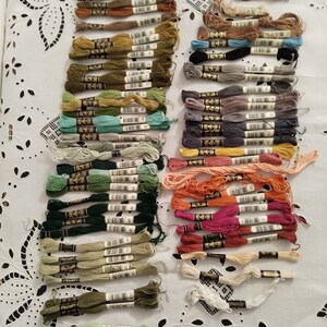 Peut inclure: Une collection de bobines de fil à broder de différentes couleurs, notamment le vert, le bleu, le rouge, l'orange, le marron et le beige. Les bobines sont disposées en rangées sur une surface blanche.