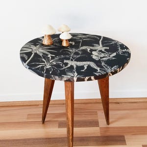 Può includere: Un tavolino rotondo con un piano nero e bianco con stampa giungla con leopardi e fogliame. Il tavolo ha tre gambe in legno ed è sormontato da tre figurine decorative a forma di fungo. Il tavolo ha un diametro di circa 61 cm.