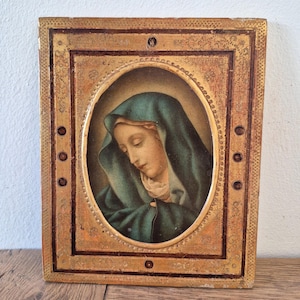 Puede incluir: Icono religioso antiguo con un retrato de una mujer con un velo verde azulado. El retrato está enmarcado en un óvalo, rodeado por un borde dorado con detalles decorativos. El marco es rectangular con un acabado envejecido.