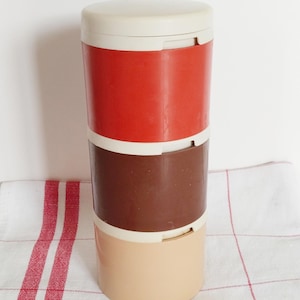 May include: A vintage, three-tiered food container with a cream-colored lid and base. The middle sections are red and brown, separated by cream-colored bands. The container is cylindrical and sits on a white and red checkered cloth.