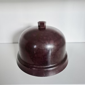 Könnte beinhalten: Eine burgunderfarbene, kuppelförmige Cloche mit strukturierter Oberfläche und einem geometrischen Knopf oben. Die Cloche steht auf einem passenden Sockel und erzeugt eine Vintage-Ästhetik. Das Design ist schlicht und elegant.