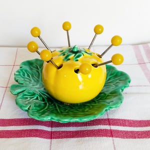 Peut inclure: Un plat en céramique jaune en forme de tomate avec huit brochettes en plastique jaunes. Le plat est posé sur une assiette en céramique verte en forme de feuille.