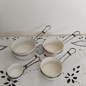 May include: Set of four white ceramic measuring cups with wire handles. The cups are nested together and have a white background with a subtle pattern.