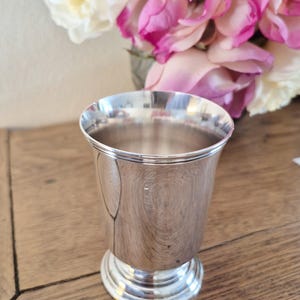 Op de afbeelding: Een zilverkleurige metalen beker met een uitlopende rand en een voet. De beker is hoogglans gepolijst en reflecteert het licht. Op de achtergrond een boeket roze en witte bloemen, wat duidt op een decoratief of verzamelobject.
