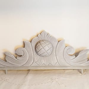 Può includere: Elemento architettonico decorativo in legno verniciato di grigio con un design centrale a sfera e volute ornate. Il pezzo è esposto su una superficie bianca, con un vaso di rose rosa e gialle a sinistra.