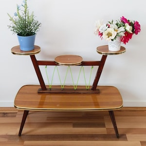 May include: A mid-century modern plant stand with three tiers and a lower shelf. The stand is made of wood with a light brown finish and gold trim. The stand has a green elastic cord running across the top. The stand is decorated with two potted plants, one with blue flowers and one with white and pink flowers.