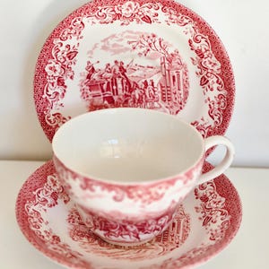 May include: A vintage tea set featuring a red and white toile pattern. The set includes a teacup, saucer, and a plate. The design depicts a rural scene with figures and buildings, surrounded by floral borders. The teacup has a handle.
