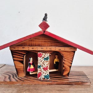 May include: A wooden music box shaped like a house with a red roof and a small black bird on top. Inside are two figurines and a decorative floral element. The house is made of wood and sits on a wooden base.