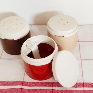 May include: Three vintage plastic containers in brown, red, and beige, each with a white lid. One container is open, revealing a white spoon. The containers are on a red and white checkered cloth.