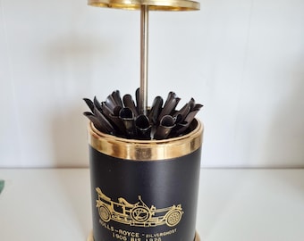 May include: A vintage black and gold Rolls-Royce Silver Ghost toothpick holder. The cylindrical holder features a gold rim and base, with a gold illustration of the car and text. The top has a gold handle and a lid.