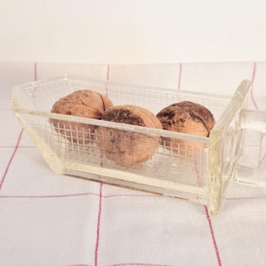 May include: A clear glass rectangular dish with a grid-patterned exterior and a single side handle. The dish contains three whole brown walnuts with textured shells. The glass has a transparent quality and a structured, geometric design.