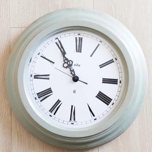 Può includere: Orologio da parete rotondo con cornice verde chiaro e quadrante bianco. L'orologio presenta numeri romani e lancette nere, con la parola "odo" stampata sul quadrante. L'ora è circa le 11:55.