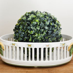 May include: White ceramic fruit bowl with a green and yellow floral design and a decorative grapevine handle. The bowl has a lattice design and is filled with a green artificial topiary.