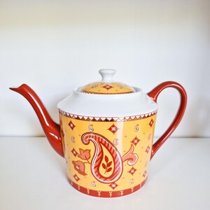 Può includere: Una teiera in ceramica con corpo giallo e motivo paisley rosso, bordo superiore bianco e coperchio abbinato. La teiera presenta un manico rosso ricurvo e un lungo beccuccio rosso, decorata con piccoli motivi a rombi e fiori di colore rosso.