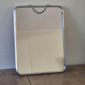 Peut inclure: Un miroir mural rectangulaire doté d'un cadre en métal argenté fin aux coins arrondis. Une petite chaîne en métal argenté délicate est fixée au sommet du cadre, permettant de suspendre le miroir verticalement sur une surface murale.