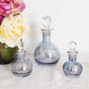 May include: Three vintage blue glass perfume bottles with crystal stoppers. The bottles have etched starburst and floral designs. The bottles are arranged on a white surface with a floral arrangement in the background.