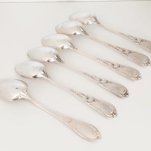 May include: Six silver spoons with decorative handles and oval bowls are arranged diagonally on a white surface. The spoons have a reflective, polished finish, suggesting they are made of sterling silver. The spoons are likely for serving.