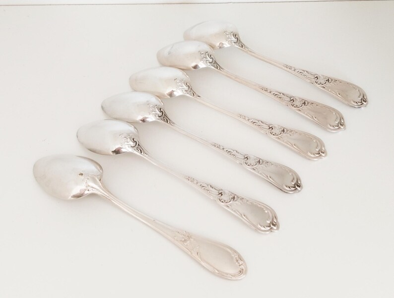 May include: Six silver spoons with decorative handles and oval bowls are arranged diagonally on a white surface. The spoons have a reflective, polished finish, suggesting they are made of sterling silver. The spoons are likely for serving.