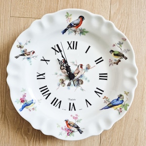 May include: White ceramic wall clock with a scalloped edge, featuring painted birds and floral accents. The clock face has black Roman numerals and black clock hands. The clock is approximately 25 cm in diameter.