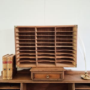 May include: A vintage wooden desk featuring a large letter sorter, a small drawer, and two antique books. The desk has a warm, brown finish and brass corner accents. A decorative feather and candle holder are also present.
