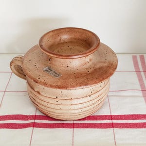 May include: A speckled, tan ceramic butter dish with a lid and handle. The dish has horizontal lines around the base and sits on a white and red striped cloth. The lid has a raised center.