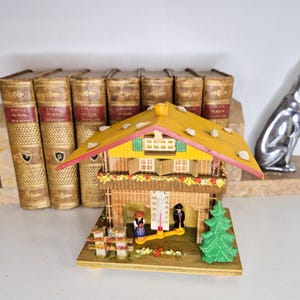 May include: A miniature wooden chalet with a yellow roof and decorative stones. The chalet features a thermometer, small figures, and a green plastic tree. Behind it are vintage books and a silver dog statue.