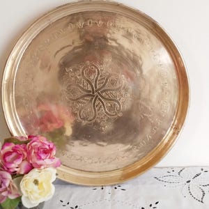 Puede incluir: Una bandeja redonda de metal dorado con un intrincado diseño floral grabado en el centro. La bandeja tiene un borde elevado y está decorada con un patrón repetitivo alrededor del borde.