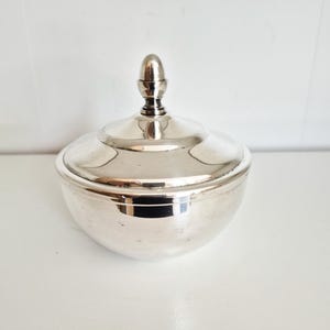 Peut inclure: Un bol argenté avec couvercle, base arrondie et petit bouton décoratif. La surface est réfléchissante et le couvercle s'ajuste parfaitement. L'objet est probablement décoratif ou pour le rangement.