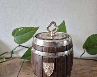 May include: A small wooden barrel-shaped container with silver-toned metal bands and a matching lid featuring a decorative handle. A silver-toned shield emblem with a lion design is attached to the front of the dark brown wood staves.
