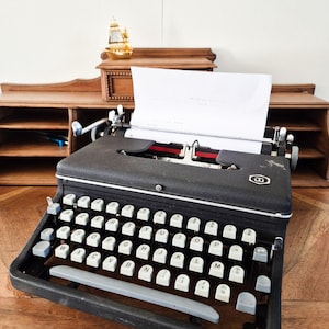 Puede incluir: Una máquina de escribir negra vintage con teclas blancas y una hoja de papel insertada. La máquina de escribir está sobre un escritorio de madera, con un pequeño barco dorado decorativo y un organizador de escritorio de madera en el fondo. Disposición QWERTY.