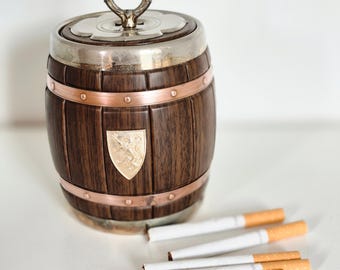 May include: A decorative wooden barrel-shaped cigarette holder with a silver lid and handle, copper bands, and a gold emblem. Several cigarettes with orange filters are scattered nearby.