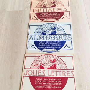 May include: A stack of vintage French paper booklets with titles like "INITIALES" and "ALPHABETS" in elegant fonts. Each booklet features illustrations of women and decorative borders. The booklets are in shades of red, blue, and peach.