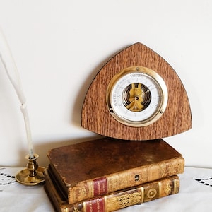 May include: A vintage wooden barometer with a brass face and a triangular wooden base. The barometer is sitting on top of a stack of three antique books.