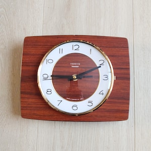 May include: A vintage wood-framed clock with a white face and black hands. The clock face has black numbers and the text "Vedette transistor" in the center. The clock is made in France.