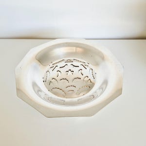 Puede incluir: Un cuenco octogonal plateado con un centro decorativo perforado. El cuenco tiene una superficie lisa y reflectante y probablemente está hecho de metal. El centro presenta un patrón de cortes curvos.