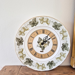 May include: A round, decorative wall clock with a white face and a gold rim. The clock features a floral design with green leaves and berries. Roman numerals mark the hours. The clock hands are black.