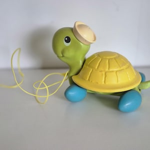 Puede incluir: Una tortuga de juguete para tirar con caparazón amarillo, cabeza verde y ruedas azules. La tortuga tiene un pequeño sombrero beige y una cuerda amarilla. El juguete está diseñado para niños pequeños.