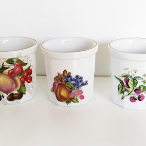 May include: Three white ceramic cups with gold rims. Each cup features a different fruit design: peaches and cherries, pears and grapes, and raspberries.