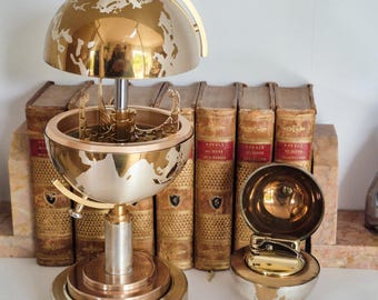 May include: A gold and silver globe-shaped music box with a hinged top, and a matching globe-shaped lighter. The music box has a detailed map design. The lighter has a round base and a hinged lid. Books are in the background.
