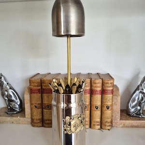 Può includere: Un portautensili in metallo color argento con un emblema decorativo. Il supporto ha una base cilindrica e un coperchio a cupola, collegati da un'asta color oro. Il supporto è pieno di utensili color oro.
