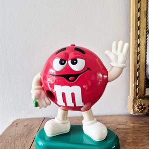 May include: A red M&M candy dispenser character with a glossy finish, white gloves, and white shoes. The figure features a large white lowercase m on its front, a wide smiling face, and is standing on a solid green oval base with a small green candy.