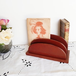 Puede incluir: Un portacartas de madera rojo con tres ranuras. El portacartas está sobre un mantel de encaje blanco con un libro titulado "Roland Encrand" y un libro antiguo encima.