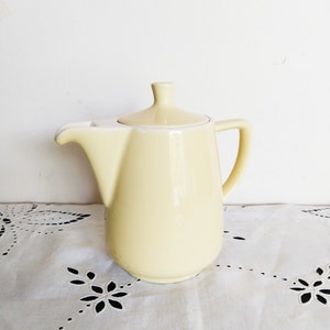 Puede incluir: Una cafetera de cerámica de color amarillo pálido con tapa y asa. La cafetera está sobre un mantel blanco con un patrón de encaje.