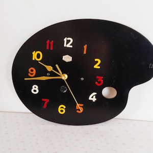 May include: A black clock shaped like an artist's palette with colorful numbers. The clock face is black with white numbers, and the numbers are in different colors, including red, yellow, and orange.
