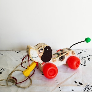 Op de afbeelding: Vintage houten trekdier hond met bruine oren, zwarte ogen en een gevlekte bruine en witte vacht. De hond heeft rode wielen en een gele trekkoord. Het speelgoed ligt op een witte kanttafelkleed.