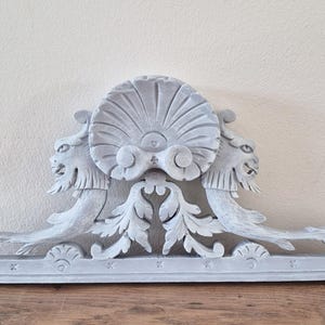 Op de afbeelding: Lichtgrijs, sierlijk architecturaal element met een schelpachtig midden en leeuwenkopdetails. Het decoratieve stuk heeft gesneden bladeren en een rechthoekige basis, perfect voor wanddecoratie of meubelversiering.