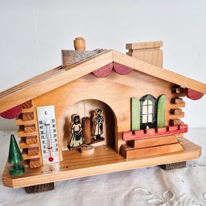 May include: Wooden chalet-style music box with a thermometer and miniature figures. The house features a red roof trim, green shutters, and a small green tree. The thermometer displays Celsius. The base is supported by small wooden blocks.