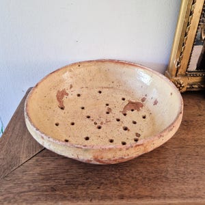 Pode incluir: Um escorredor de cerâmica redondo e raso com um interior esmaltado na cor creme, apresentando uma textura salpicada e desgaste visível. A tigela possui numerosos orifícios de drenagem na base e uma borda espessa de terracota sem esmalte.